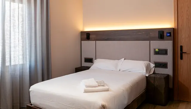 Habitación doble con cama grande vestida de blanco, cabecero de madera y tela iluminado por luz LED, dos mesitas