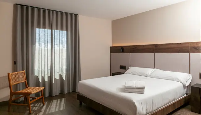 Habitación doble con cama grande vestida de blanco, cabecero de madera y paneles tapizados, silla de mimbre