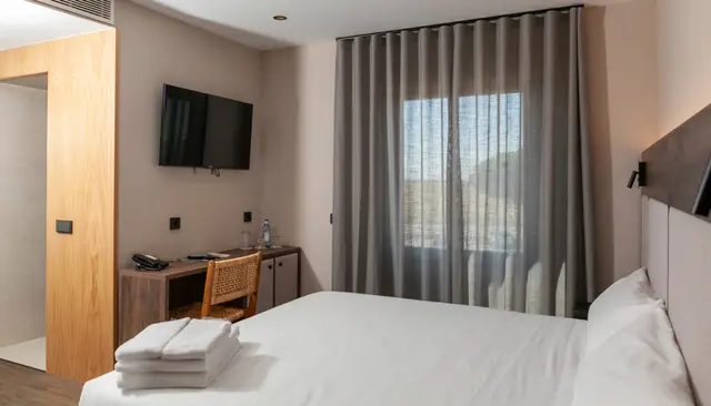 Vista de una habitación doble con cama grande vestida de blanco, toallas dobladas, escritorio con silla, televisor