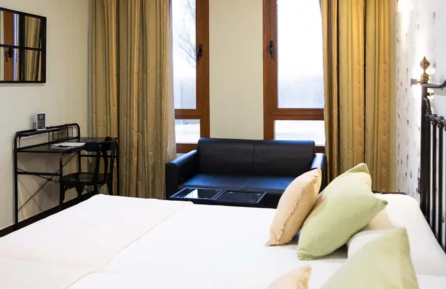 Vista de una suite con cama grande en primer plano con almohadas blancas y verdes, sofá negro junto a una ventana
