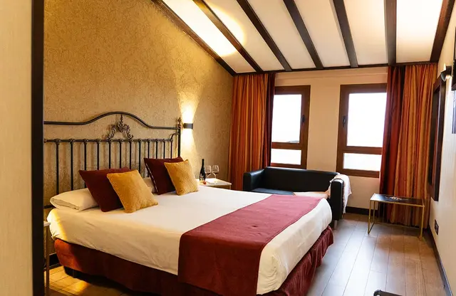 Vista de una suite con cama doble grande vestida con sábanas blancas, colcha roja y cojines granates y dorados.