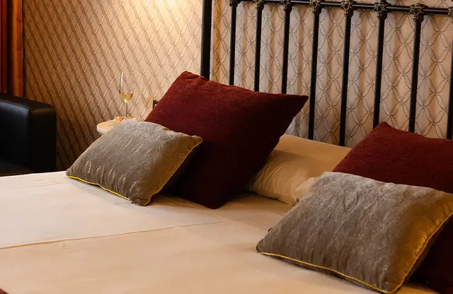 Cama matrimonial con sábanas blancas, varios cojines en tonos burdeos y grises con ribetes dorados, cabecero de forja