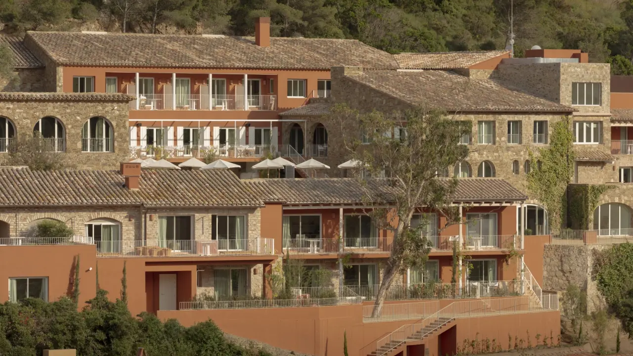 Edificio de suites con fachada de piedra y terracota, balcones y terrazas con vistas a la vegetación de la colina.