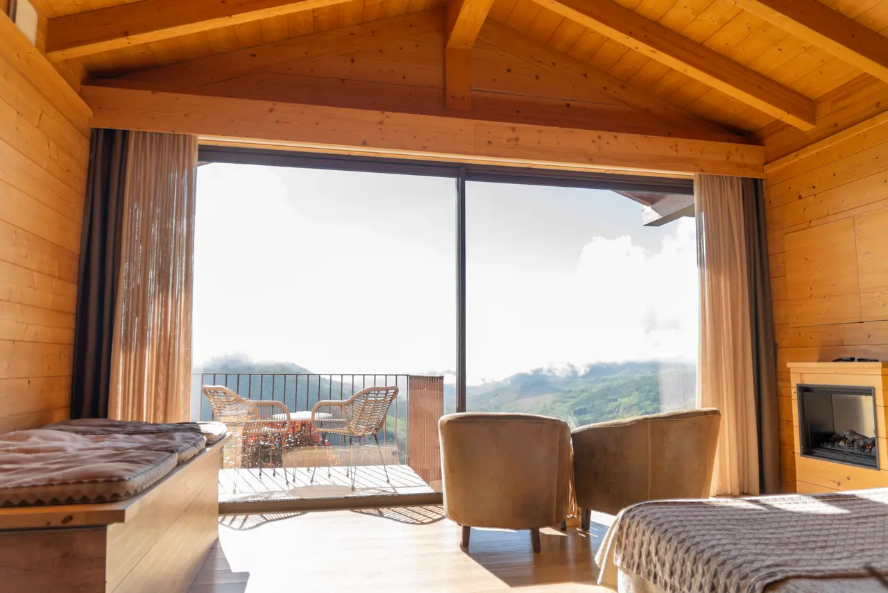 Interior de una suite con paredes y techo de madera, grandes ventanales que dan a una terraza con vistas a montañas