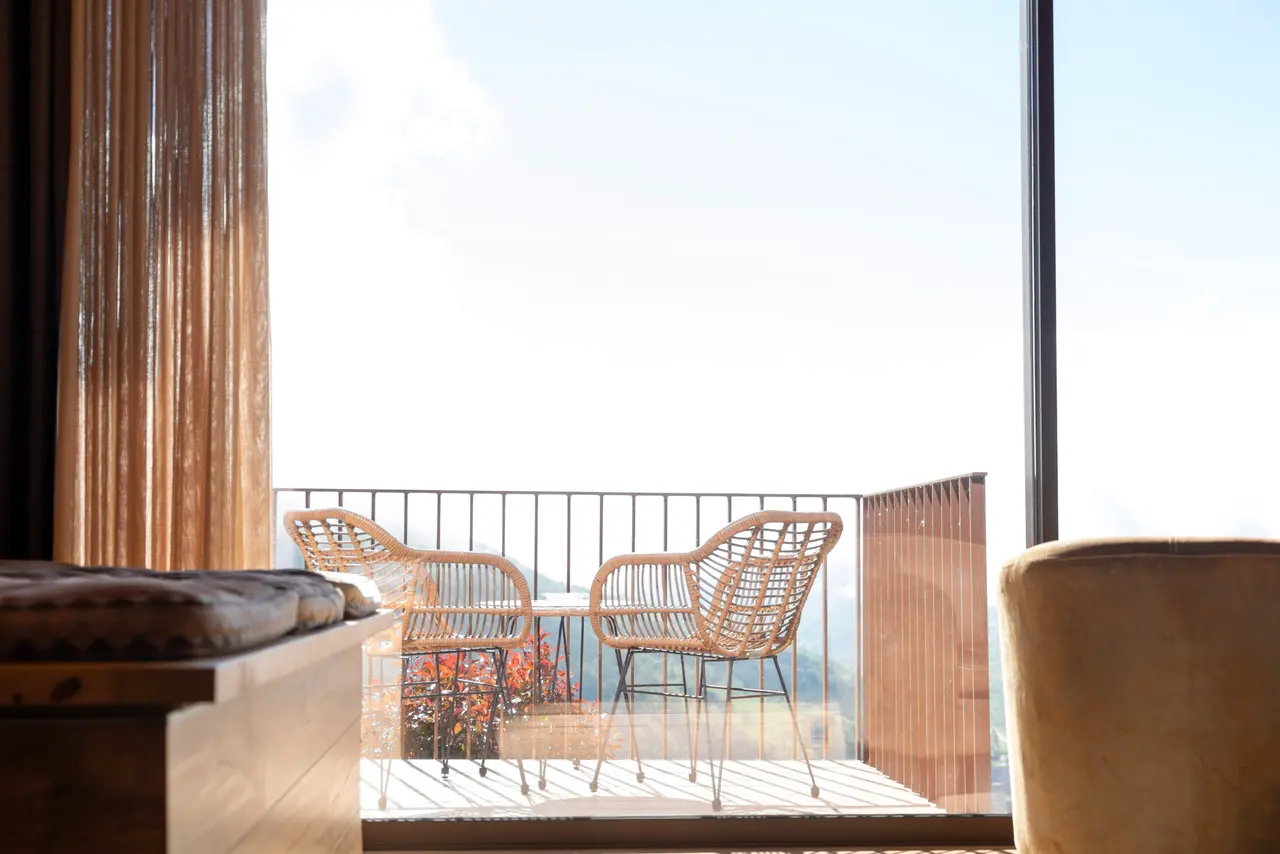 Vista desde el interior de la suite hacia una terraza con dos sillas de mimbre y una mesa, con cortinas a la izquierda