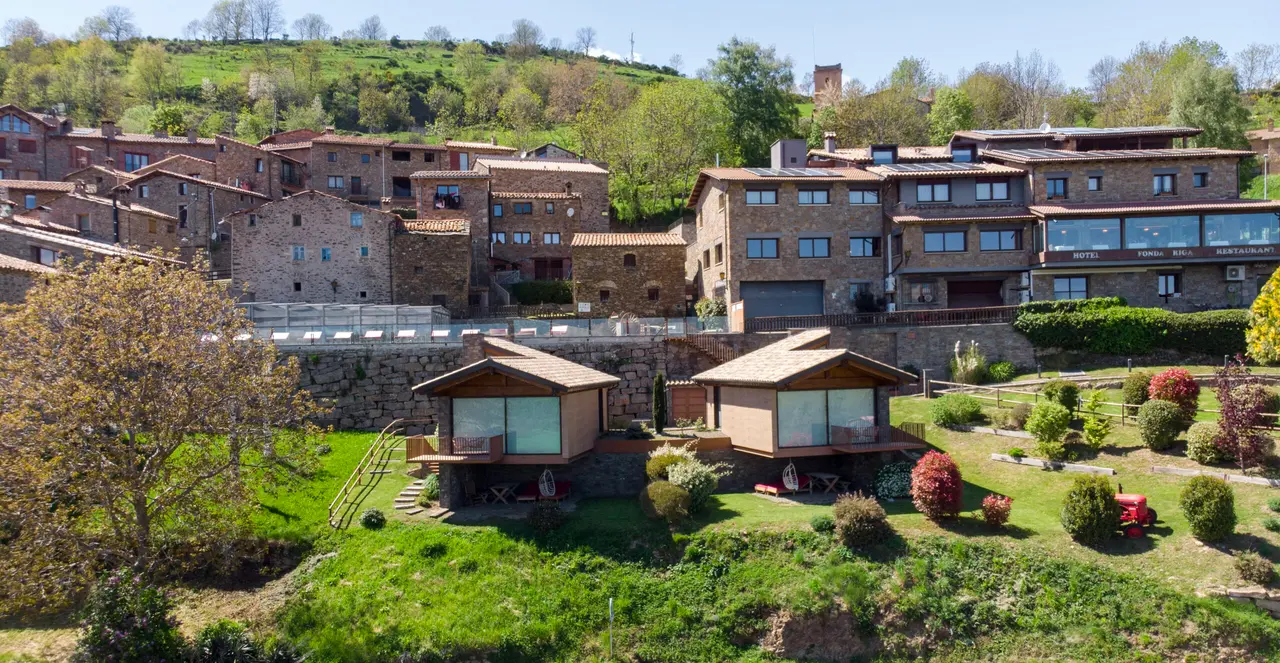 Vista exterior de dos cabañas de madera con terraza, situadas en un jardín verde con arbustos y flores
