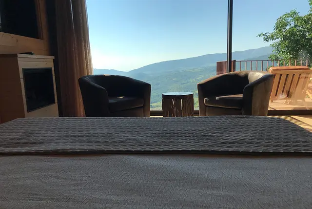 Vista desde el interior de la cabaña hacia un balcón con dos sillones oscuros y una mesa de tronco, con montañas