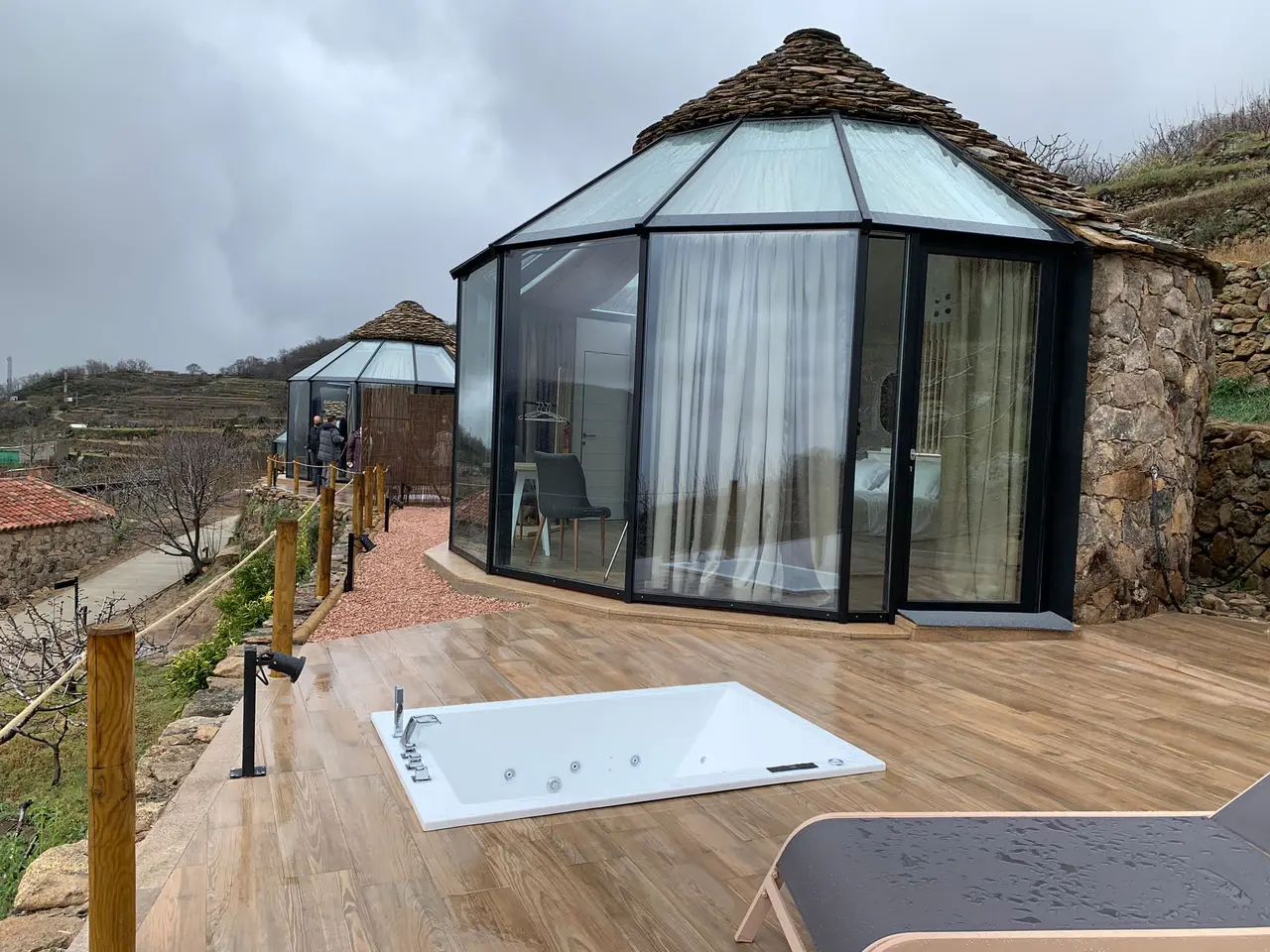 Jacuzzi blanco rectangular en terraza de madera exterior junto a chozo panorámico de cristal y piedra.