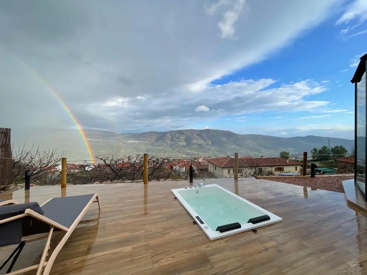 Jacuzzi exterior en terraza de madera con vistas a montañas, arcoíris y valle nublado.