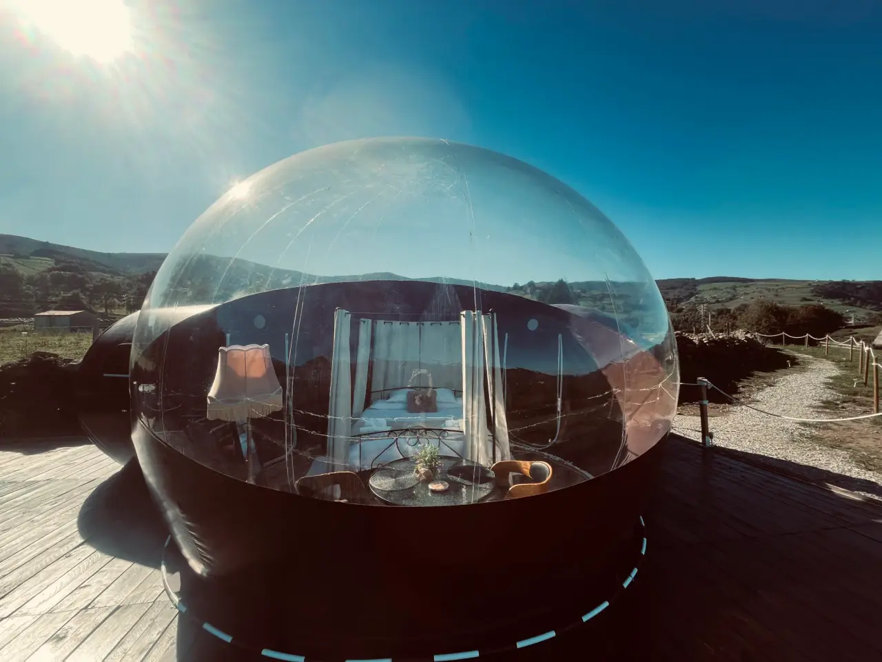 Vista exterior de una cabaña burbuja transparente sobre una terraza de madera, con interior visible que incluye cama