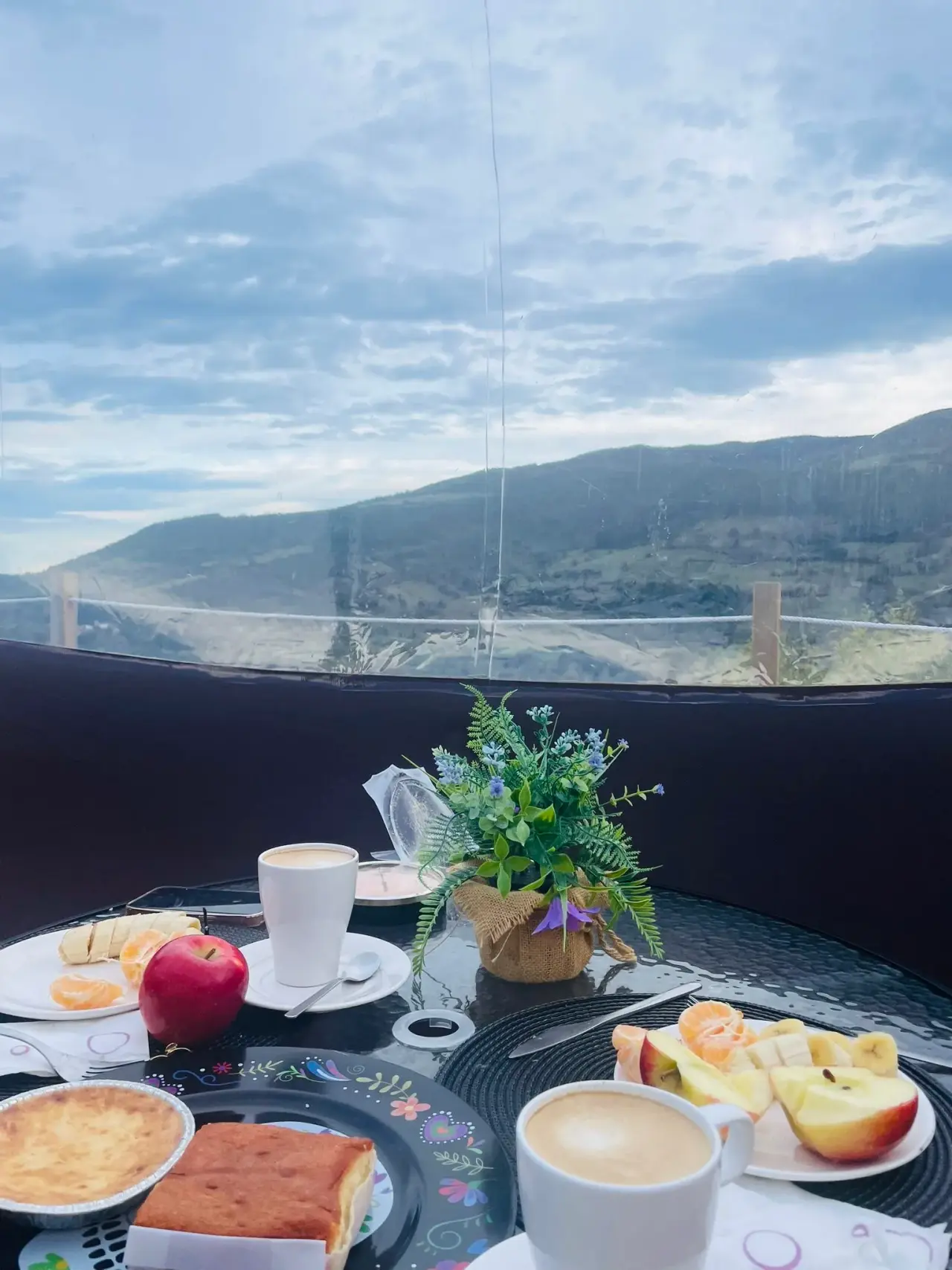 Vista panorámica de montañas verdes y nubladas desde el interior de una cabaña, con una mesa redonda en primer plano