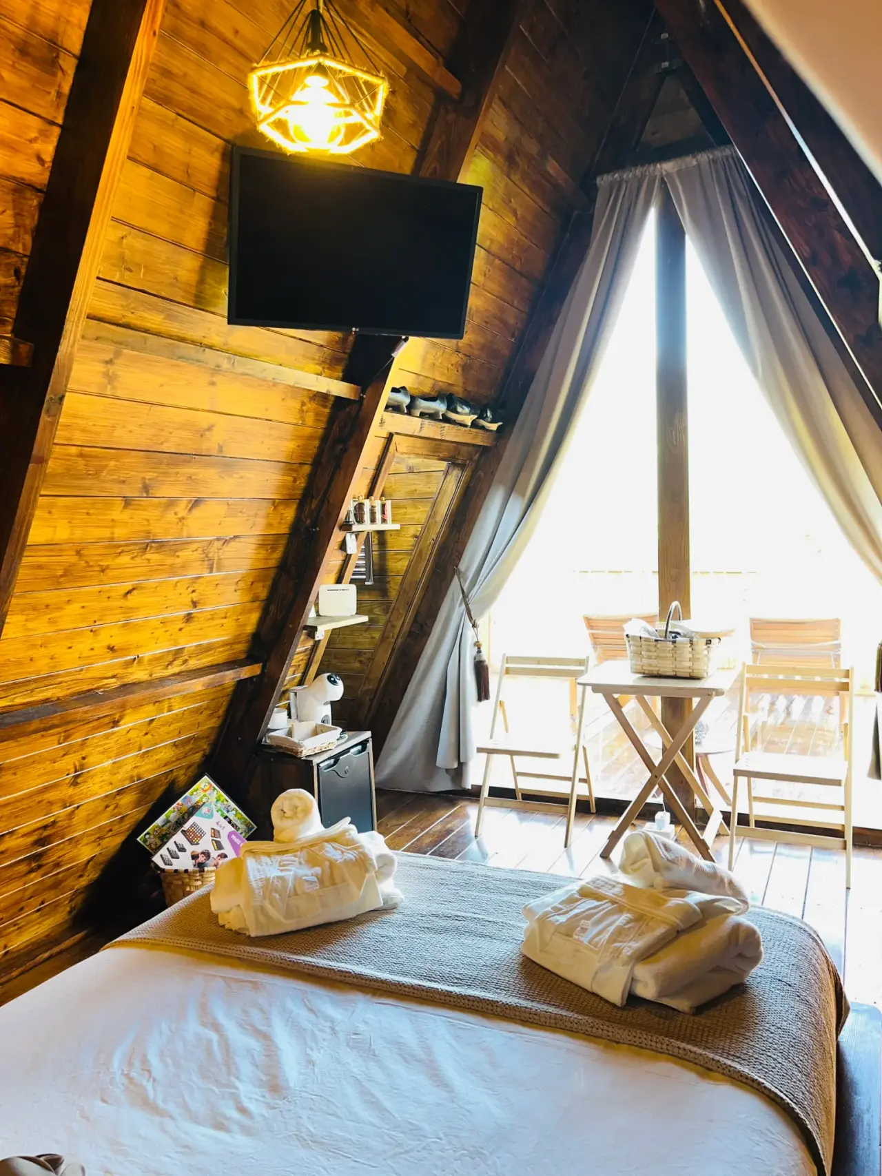 Interior de una cabaña con paredes y techo de madera, cama grande con toallas blancas dobladas, televisor montado