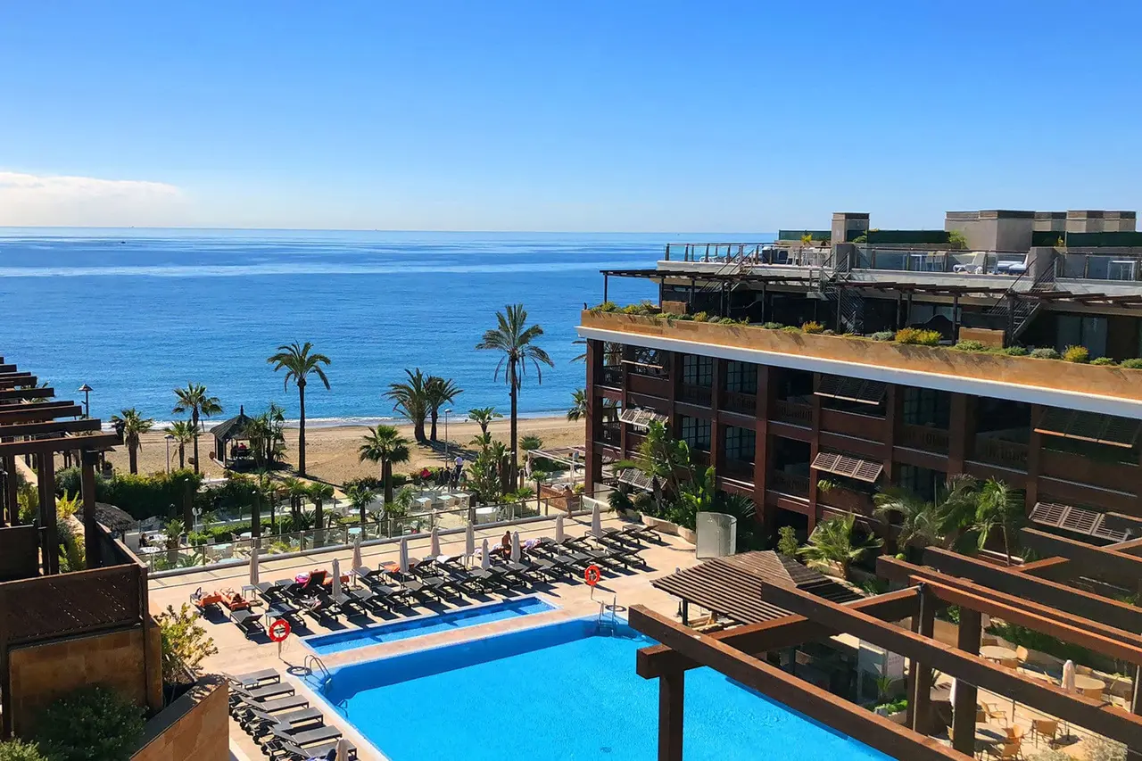 Vista panorámica desde un ático con piscina rectangular en primer plano, tumbonas, palmeras, playa de arena y el mar
