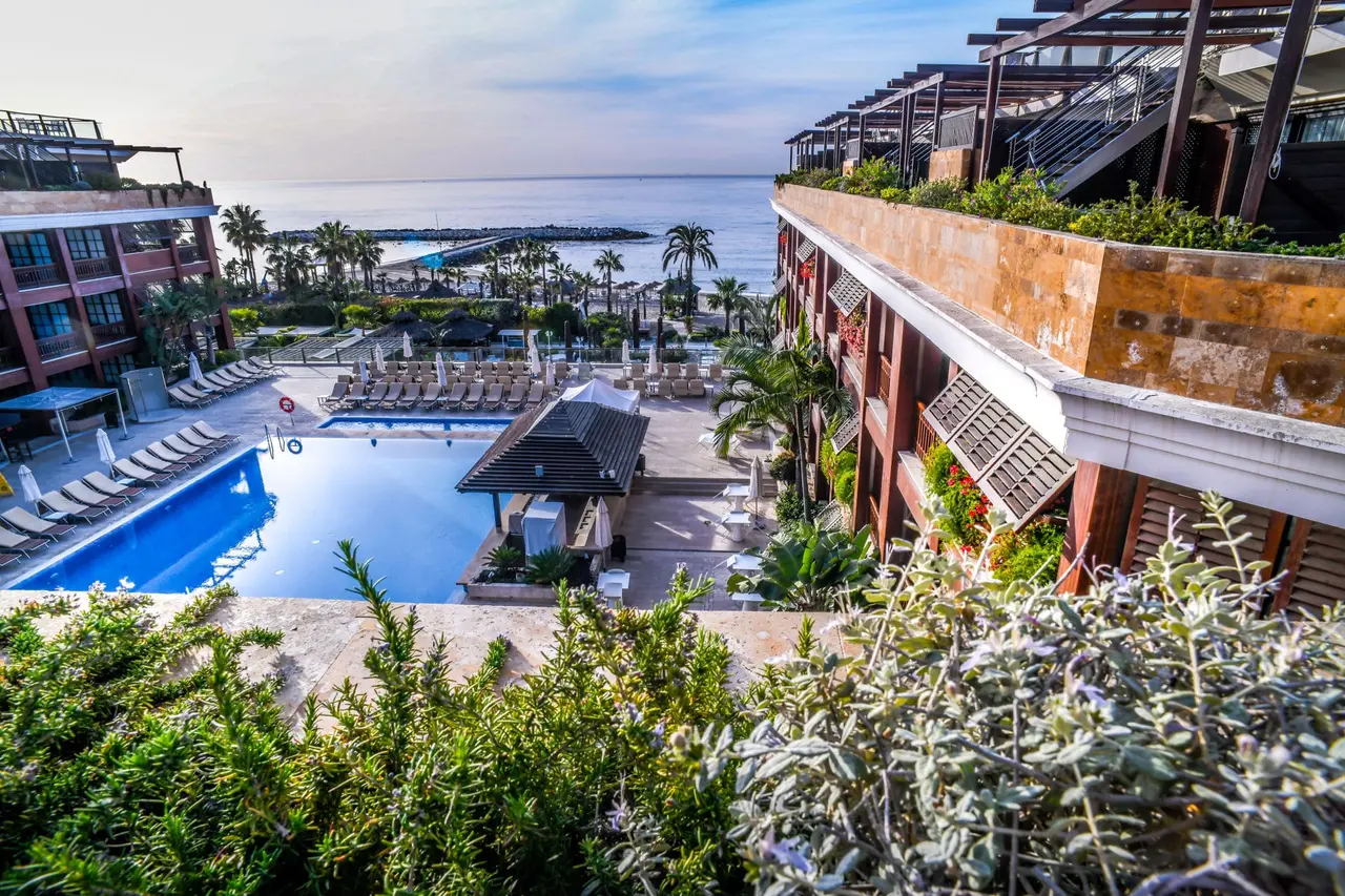 Vista exterior de la zona de piscinas y terrazas del hotel con el mar al fondo, tomada desde un ático con vegetación
