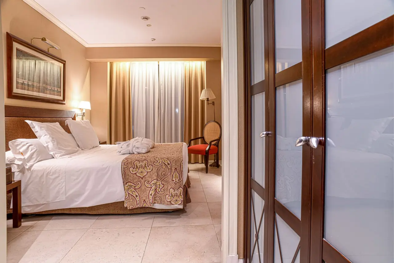 Vista de una habitación de hotel con una cama grande vestida de blanco, un banco decorativo a los pies, una silla