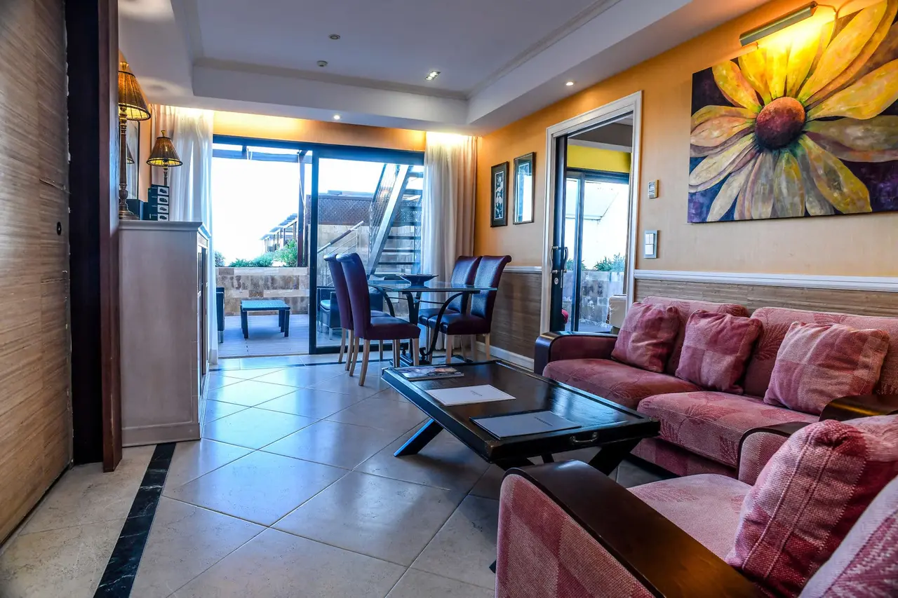 Interior de una suite con salón amueblado con sofá rosa, mesa de centro, comedor con mesa y sillas, y acceso