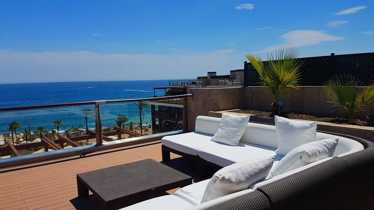 Terraza exterior con sofá blanco, mesa baja y vistas al mar bajo un cielo azul despejado.
