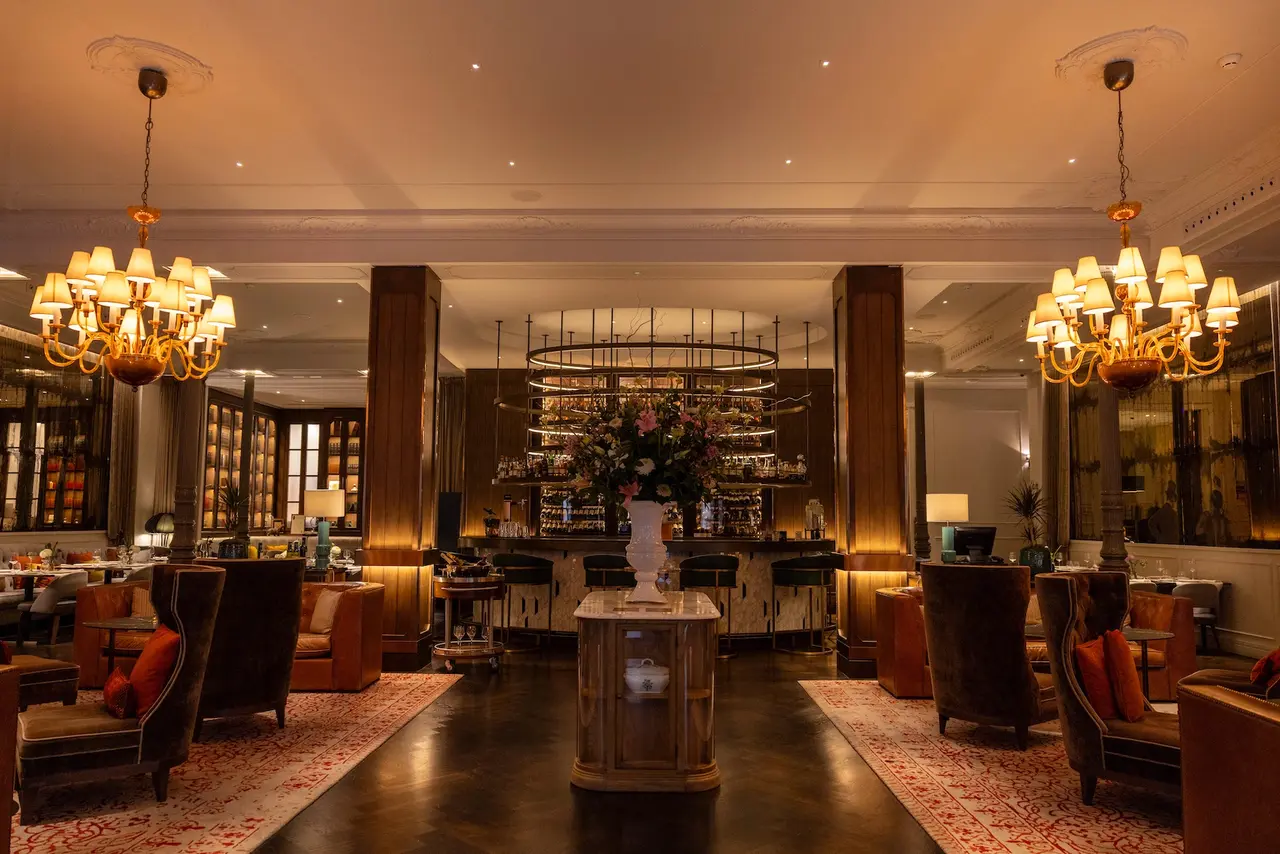 Interior de un salón elegante con techos altos, dos grandes lámparas de araña doradas, sillones de cuero marrón