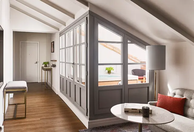 Vista interior de una suite con suelo de madera, sofá gris con cojín rojo y mesa de centro redonda.