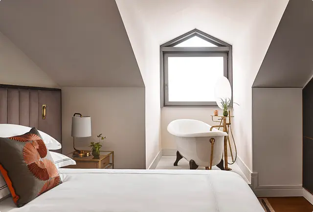 Interior de una suite con cama grande en primer plano, cabecero acolchado, mesita de noche con lámpara y planta