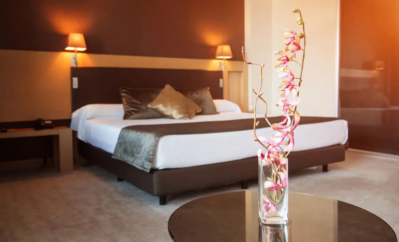 Habitación con cama grande vestida de blanco y marrón, cabecero iluminado con dos lámparas de pared, mesita de noche
