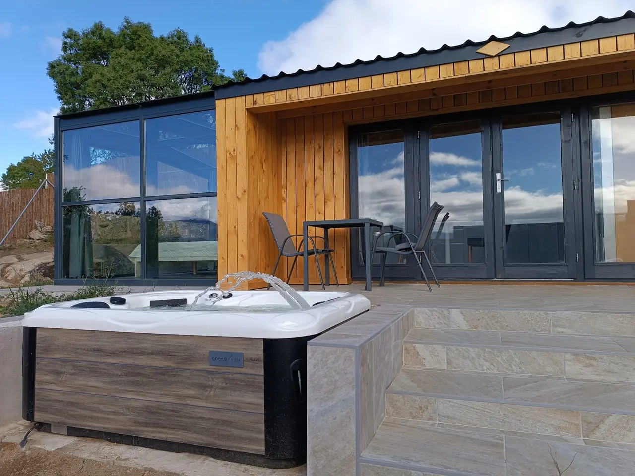 Bañera de hidromasaje exterior climatizada en la terraza de una cabaña de madera con mesas y sillas.