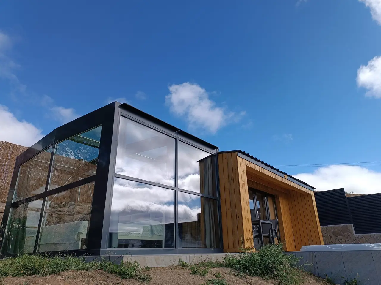 Estructura moderna de vidrio y madera con cubierta negra bajo un cielo azul con nubes, rodeada de vegetación baja