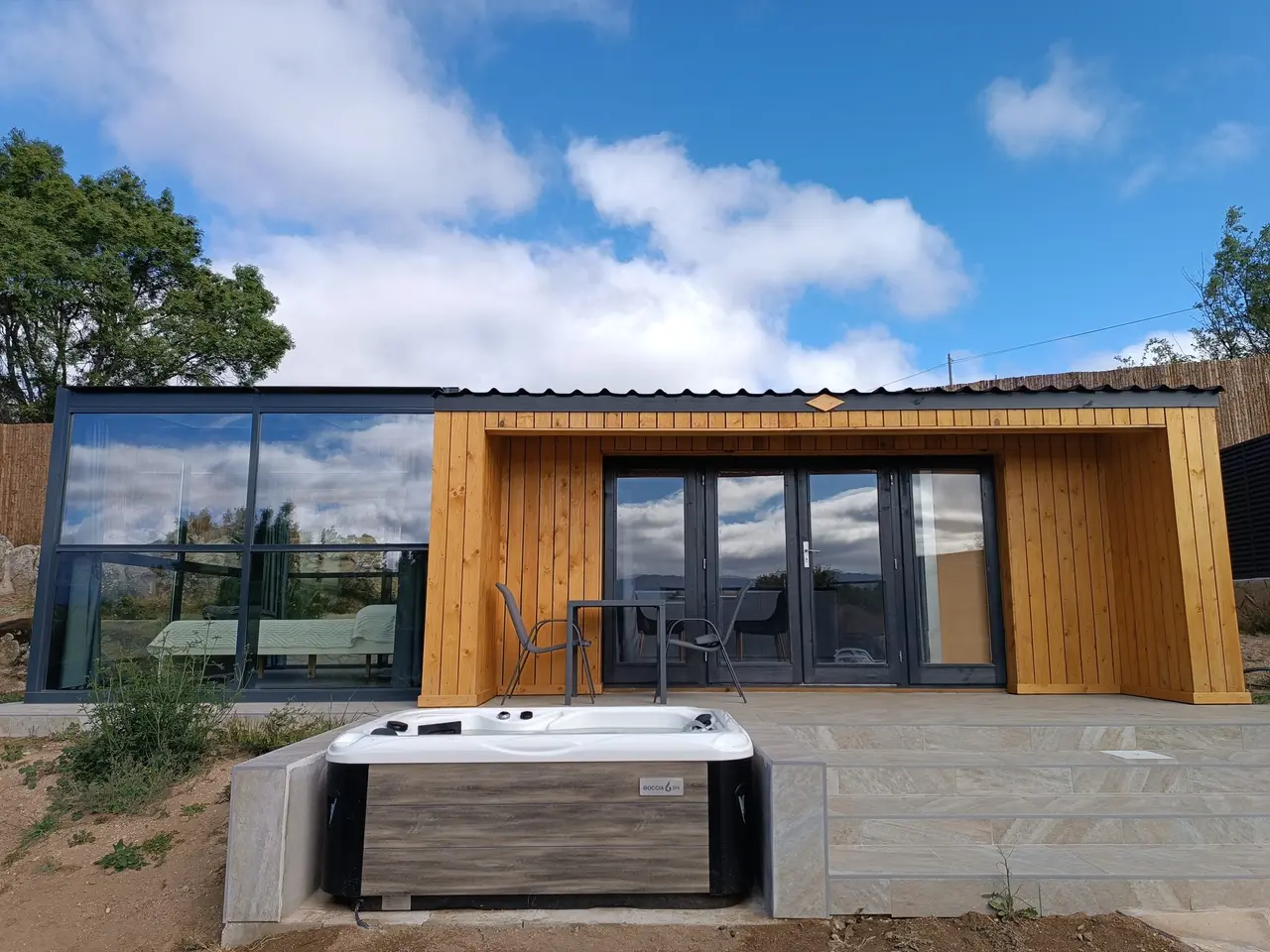 Bañera de hidromasaje exterior climatizada instalada en la terraza de una cabaña de madera con grandes ventanales.