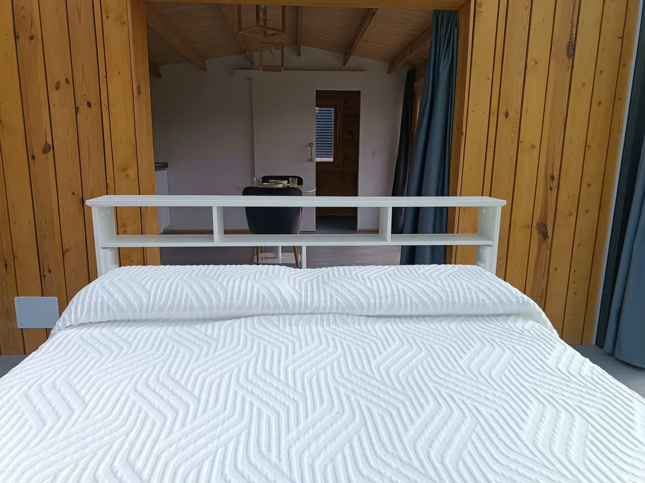 Cama con colcha blanca en habitación de cabaña con paredes de madera y vista al salón con mesa y silla.