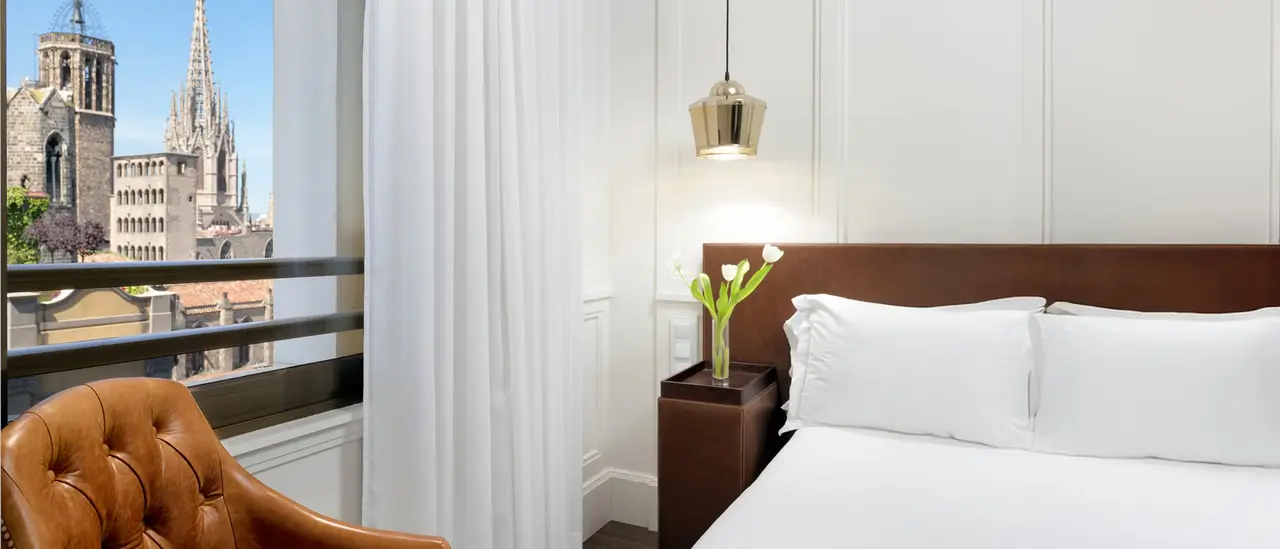 Vista de una junior suite con cama grande vestida de blanco, cabecero de madera oscura, mesita de noche con jarrón