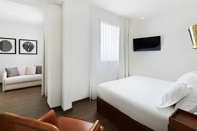 Vista de una junior suite con cama grande de matrimonio, butaca de cuero marrón en primer plano, sofá en la sala