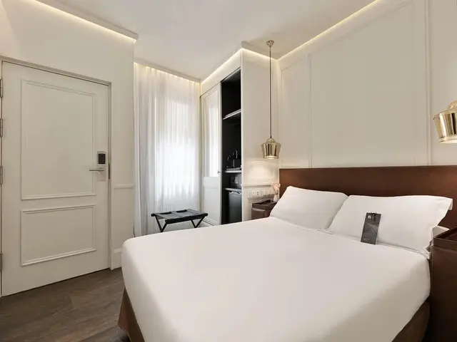 Vista de una junior suite con cama grande vestida de blanco, cabecero de madera, mesita de noche, puerta blanca