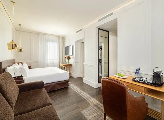 Vista de una junior suite con cama grande, sofá marrón en primer plano, escritorio de madera con silla de cuero, suelo