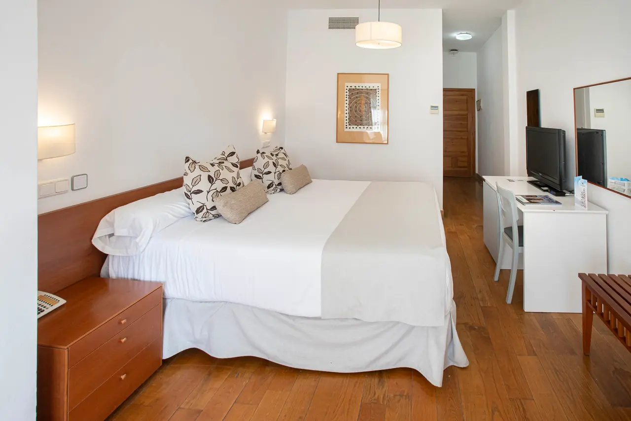 Habitación doble con cama grande vestida de blanco, escritorio con TV, mesita de noche y suelo de madera.