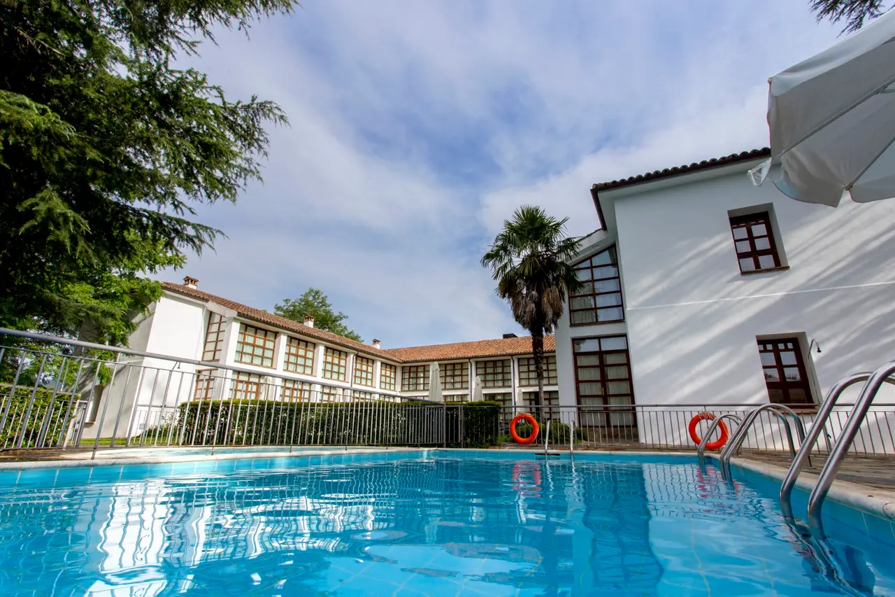 Piscina exterior rectangular con barandilla metálica y escaleras, rodeada de edificios blancos y vegetación bajo
