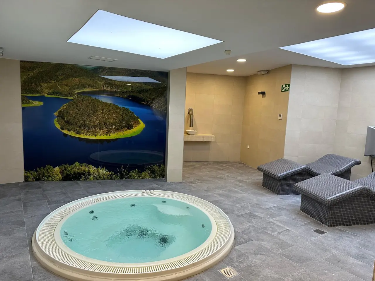 Jacuzzi circular con agua en una sala con mural de paisaje natural, tumbonas y techo con tragaluz.