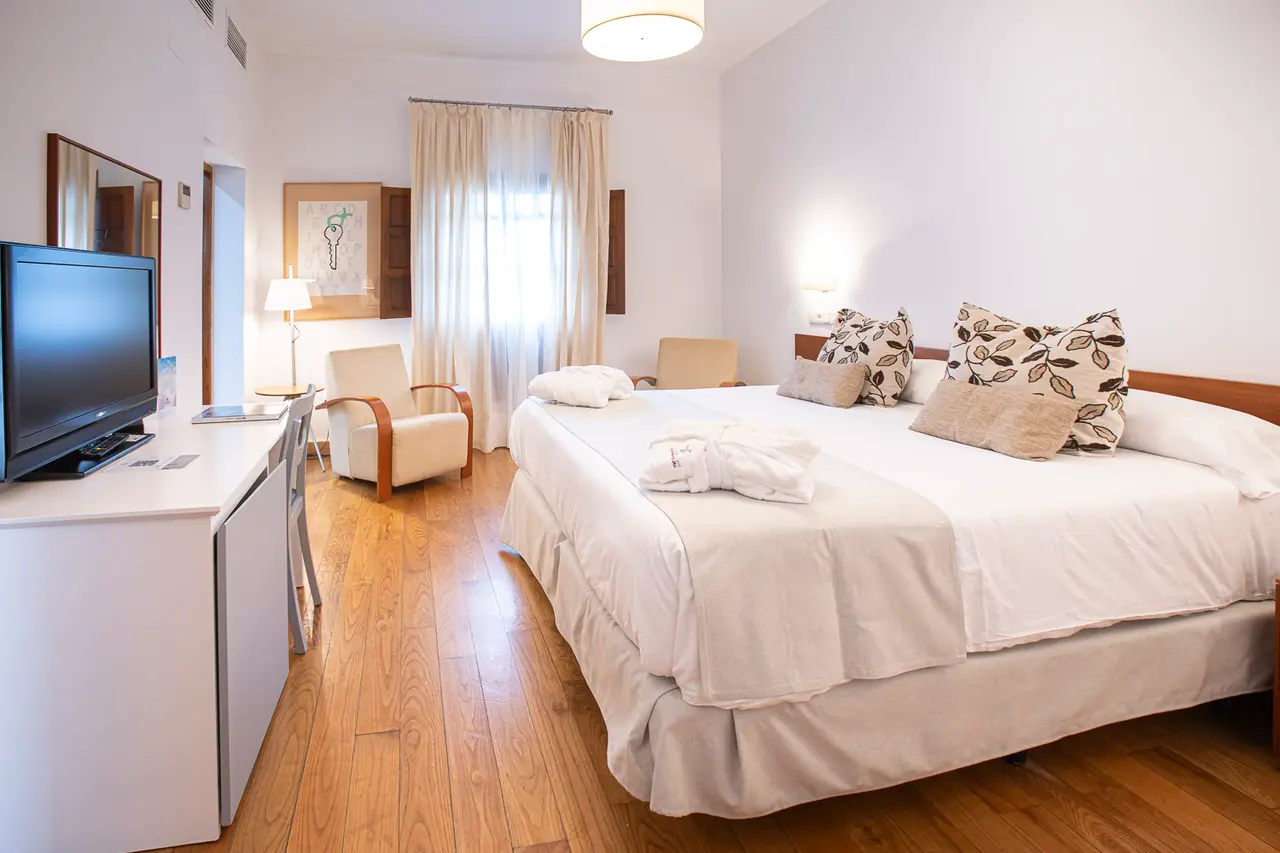 Habitación doble con cama grande vestida de blanco, escritorio con televisor, sillón y ventana con cortinas.