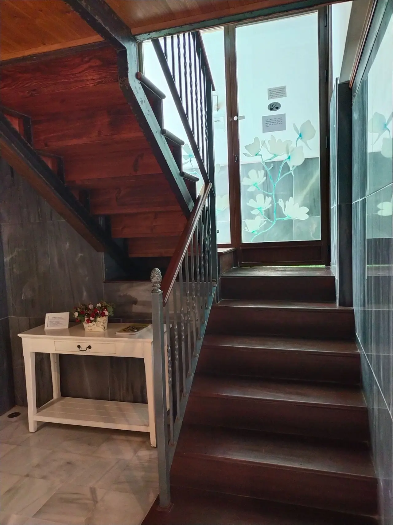 Escalera de madera oscura con barandilla de metal que conduce a una puerta de vidrio con decoración floral, al lado