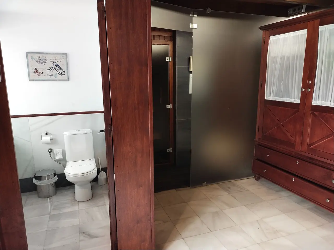 Vista de un baño con inodoro blanco, papel higiénico, cubo de basura metálico y puerta de madera que da a un espacio