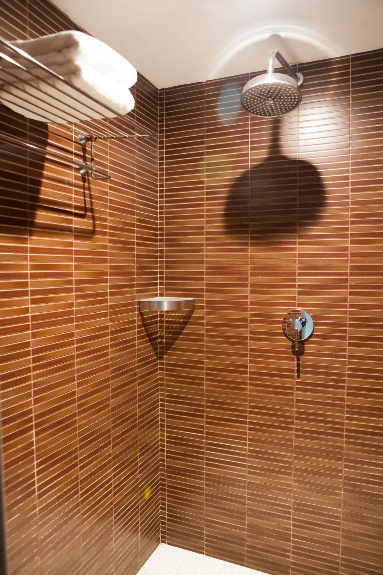 Ducha de esquina con paredes de azulejos marrones verticales, alcachofa de ducha redonda, grifo empotrado, repisa