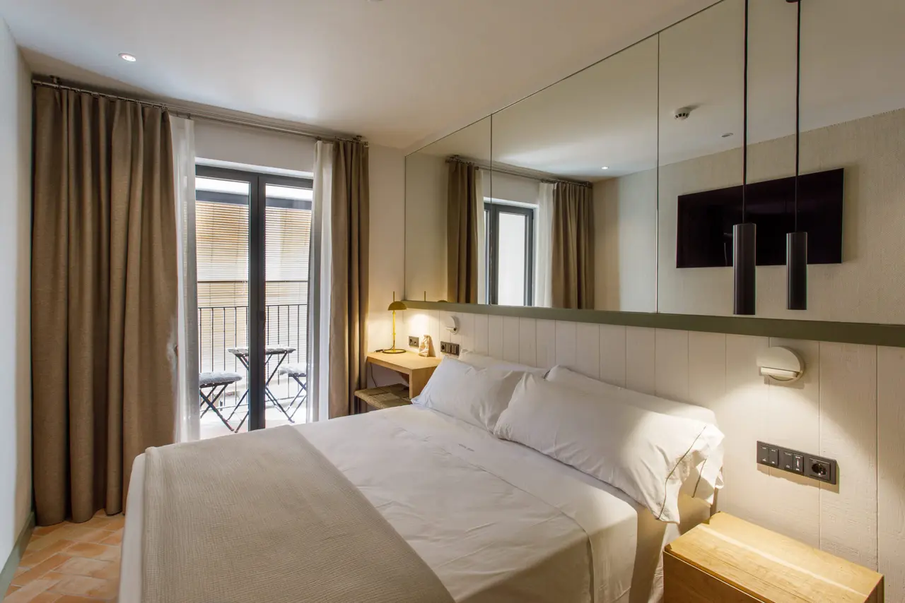 Habitación con cama doble vestida de blanco, cabecero con paneles claros y gran espejo horizontal, mesita de noche