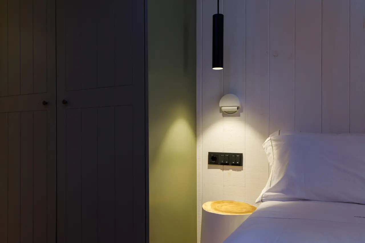 Vista de una habitación con pared de madera blanca, cama con almohadas blancas, mesita de noche redonda con luz
