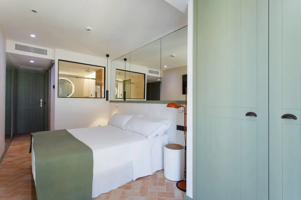 Habitación con cama doble vestida de blanco y verde, armario de puertas verdes, suelo de terracota y vistas a un baño