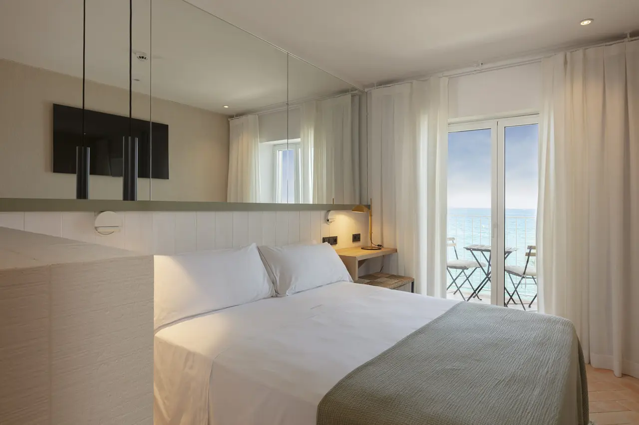 Habitación con cama doble vestida de blanco, cabecero con espejo grande, escritorio con lámpara y puertas de vidrio