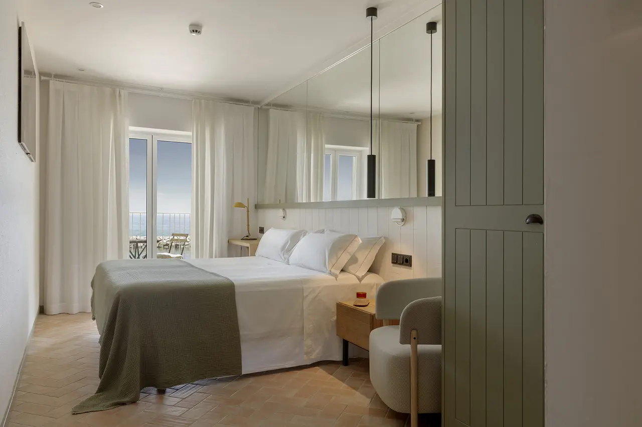 Habitación con cama doble vestida de blanco y verde, cabecero con espejo grande, dos mesitas de noche, butaca beige