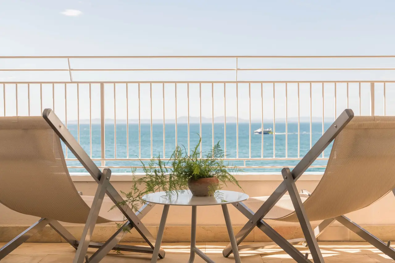 Dos sillas plegables y una mesa con planta en una terraza con barandilla frente al mar y un barco en el horizonte.