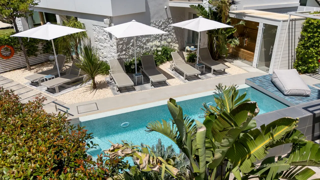 Piscina rectangular con tumbonas y sombrillas en un patio rodeado de vegetación y edificios modernos.