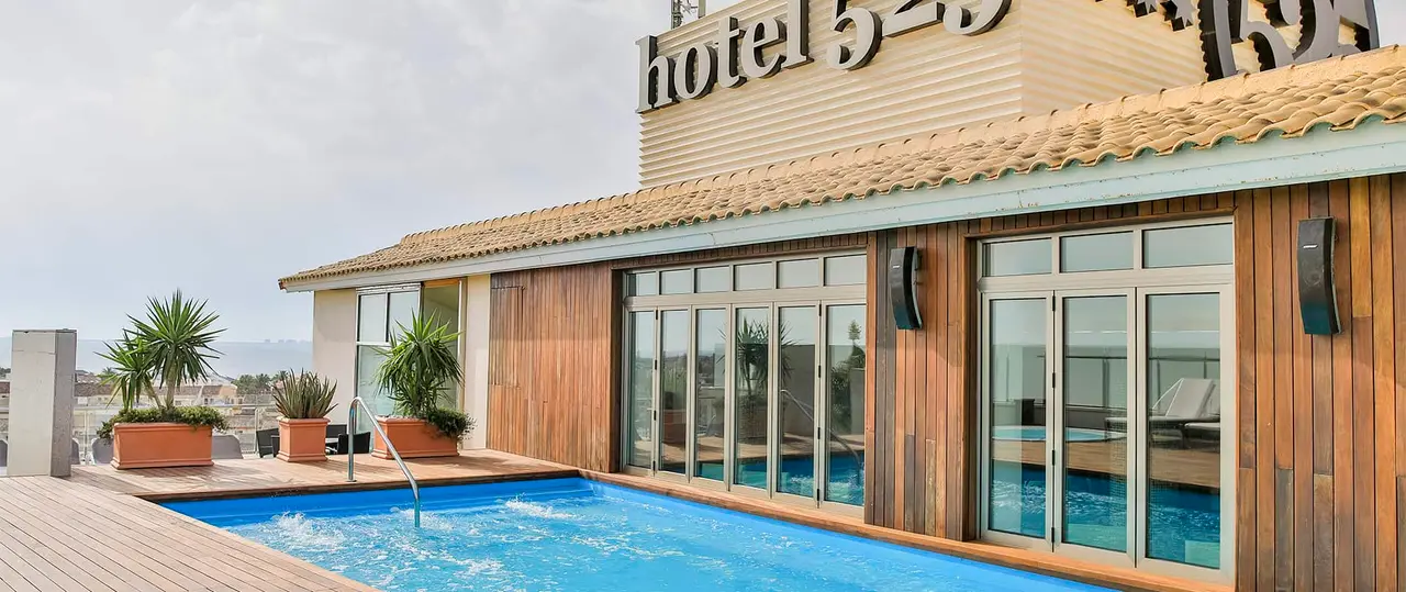 Vista de una piscina rectangular con agua azul en una terraza exterior de un hotel, rodeada de deck de madera, macetas