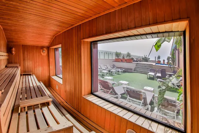 Interior de sauna de madera con bancos y ventana grande que da a una terraza exterior con tumbonas y piscina.