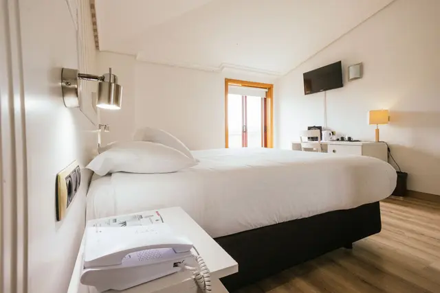 Habitación de hotel con cama doble grande vestida de blanco, mesita de noche con teléfono y lámpara de pared